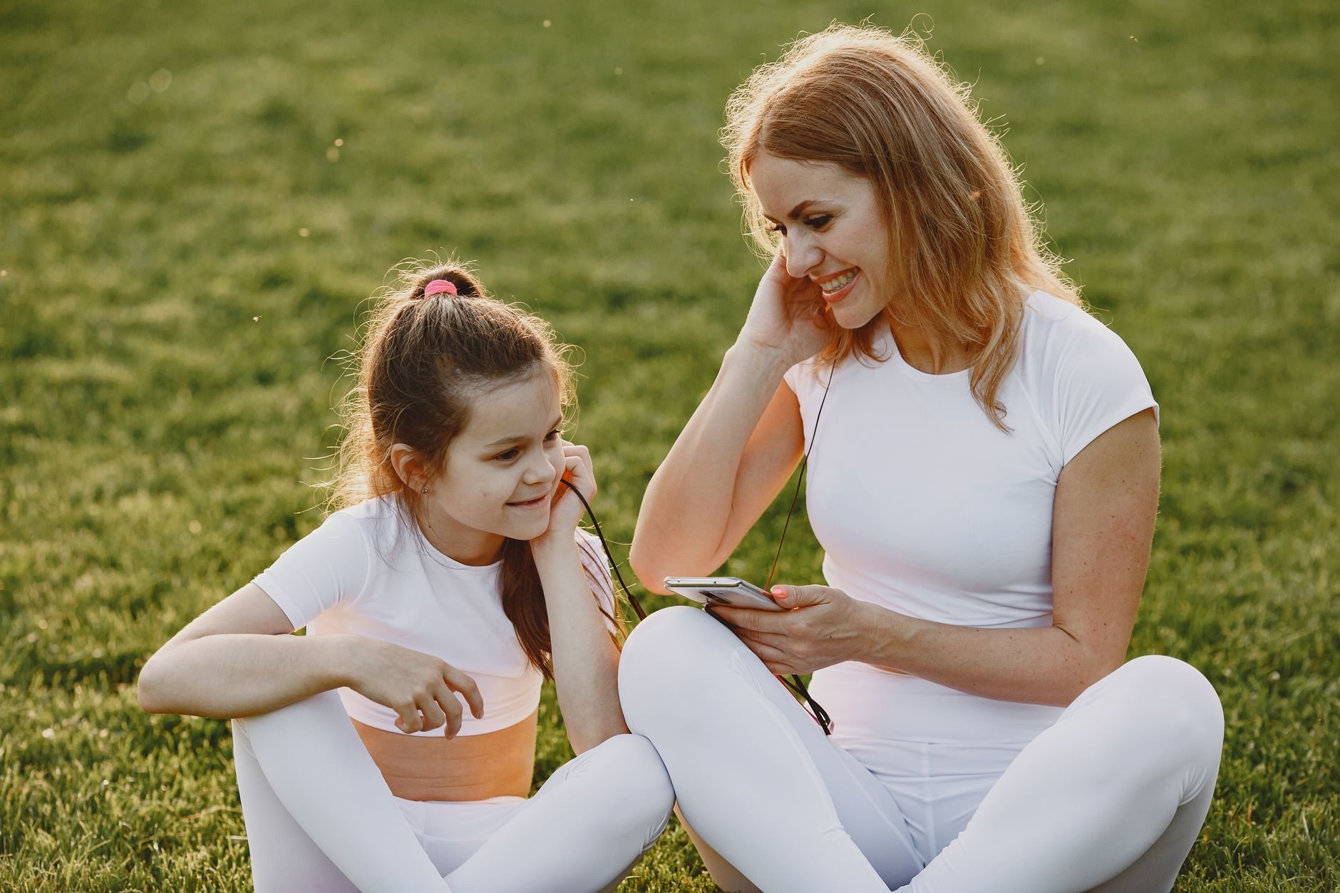 A mother and daughter sitting on grass, sharing headphones and enjoying music outdoors.
